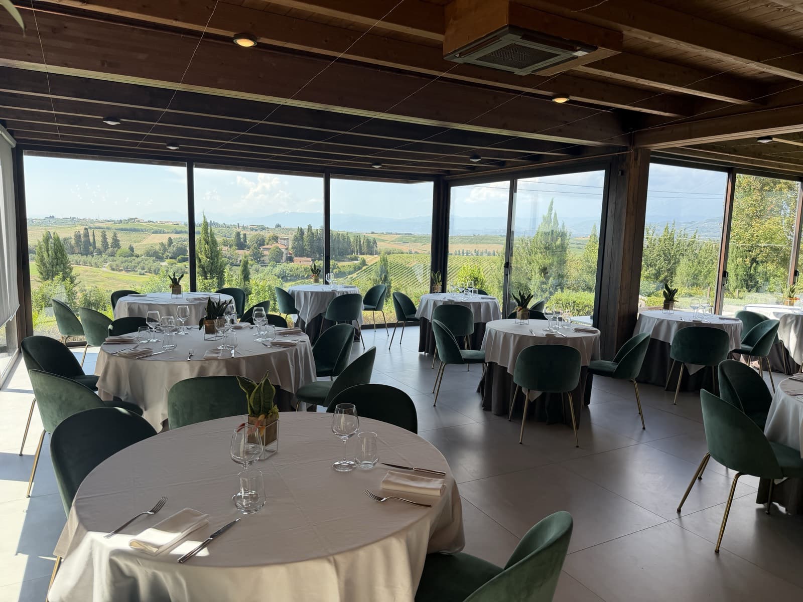The full dining room, several round tables laid in white with green chairs, panoramic windows along the far wall looking onto the valley