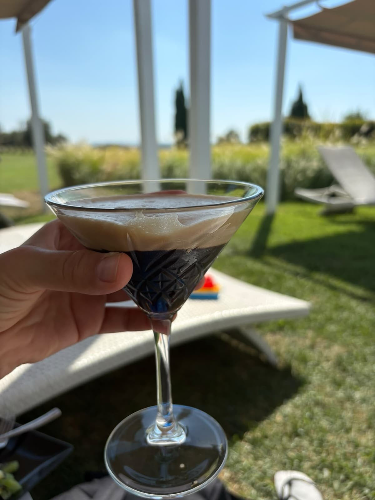 A hand holding a coupe glass of iced coffee at the poolside, with sunbeds, lawn and a white pergola behind