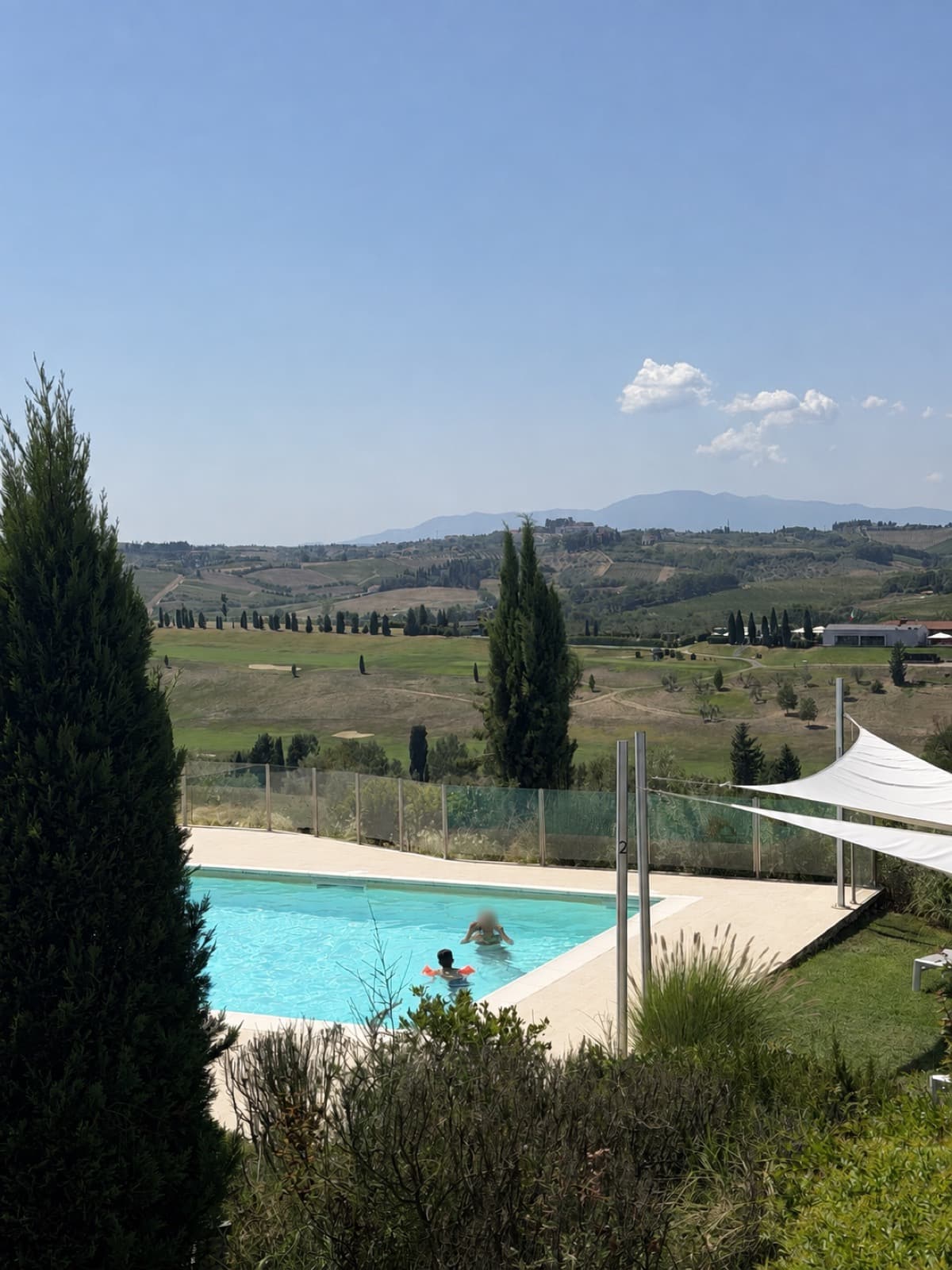 The saltwater pool seen from above between cypresses, two swimmers in the water, shade sails to one side and vineyards and hills opening beyond