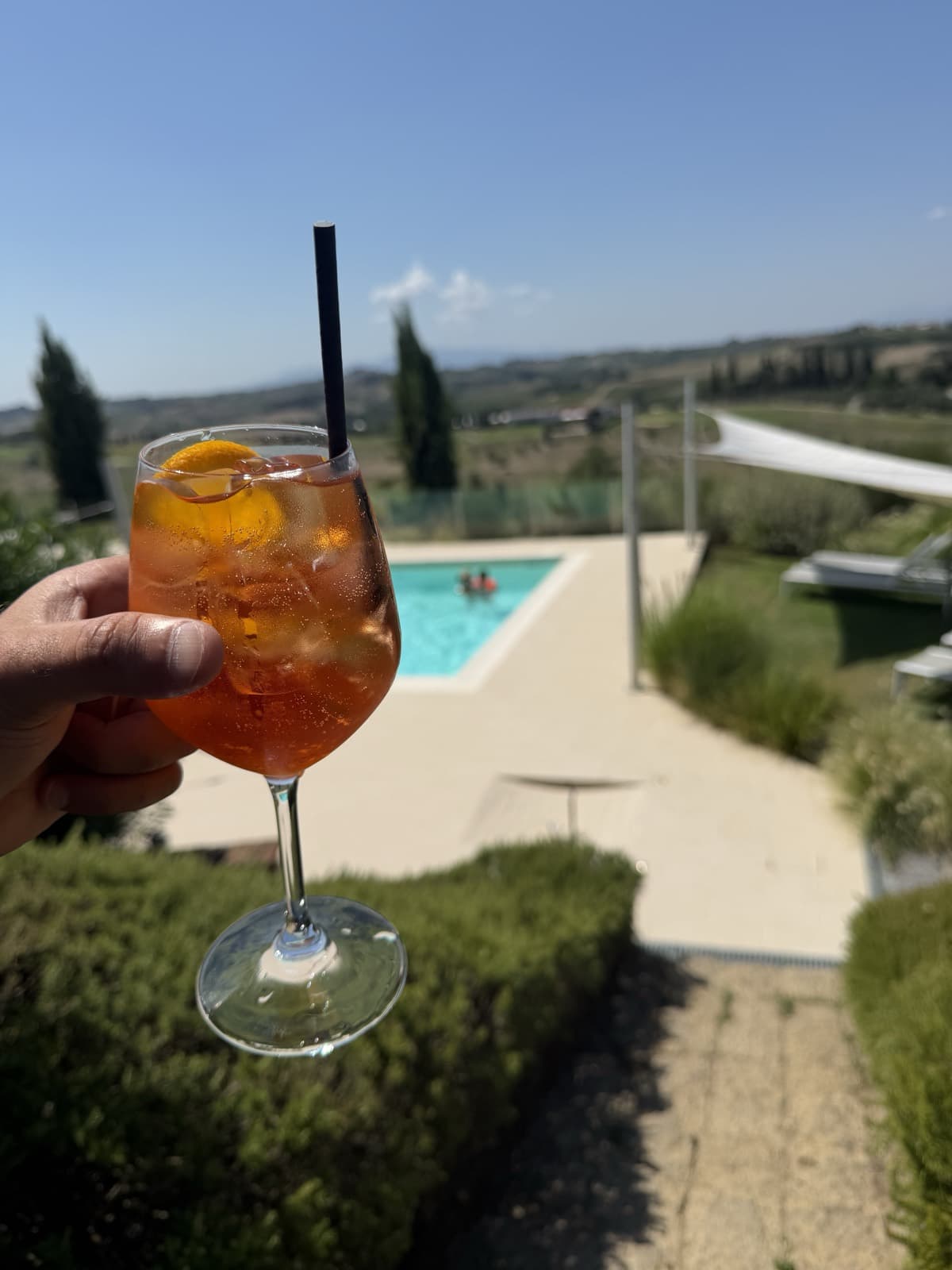 A spritz held up in one hand, orange and full of ice with a slice of orange and a black straw, the pool and its lawn below and vineyards and cypresses running to the hills behind