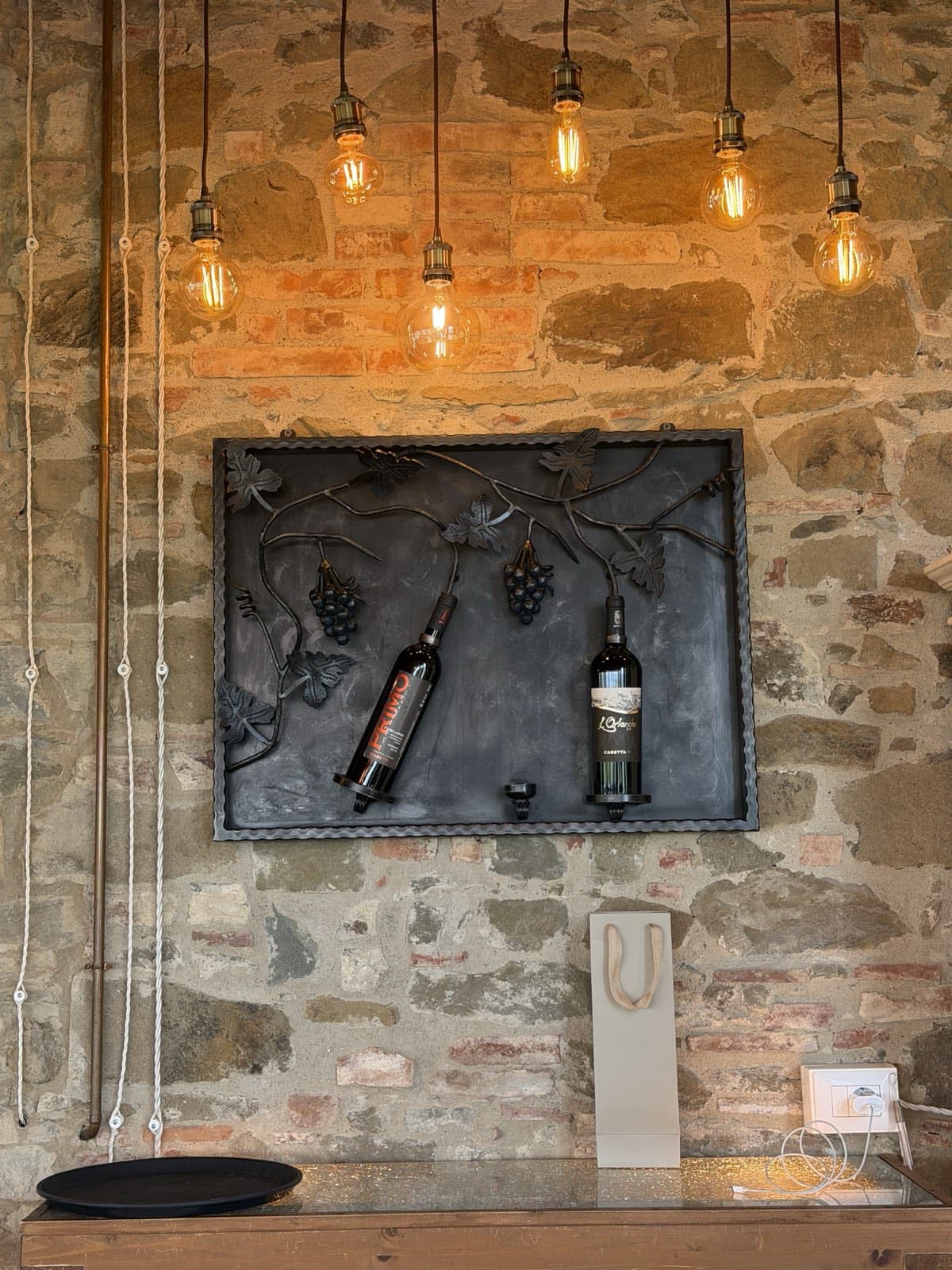 Two of the estate's bottles mounted on an ironwork panel of vines and grapes against a stone wall, filament bulbs hanging above