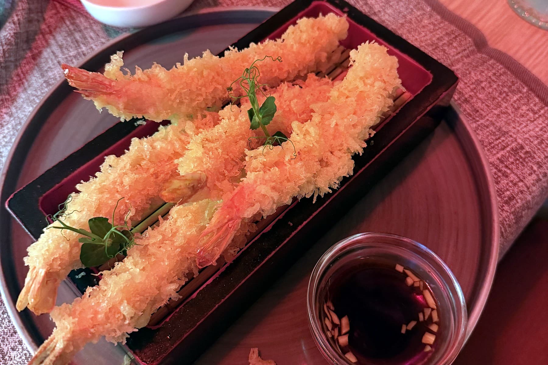 Four ebi tempura prawns in golden batter on a black tray, with a dipping sauce alongside