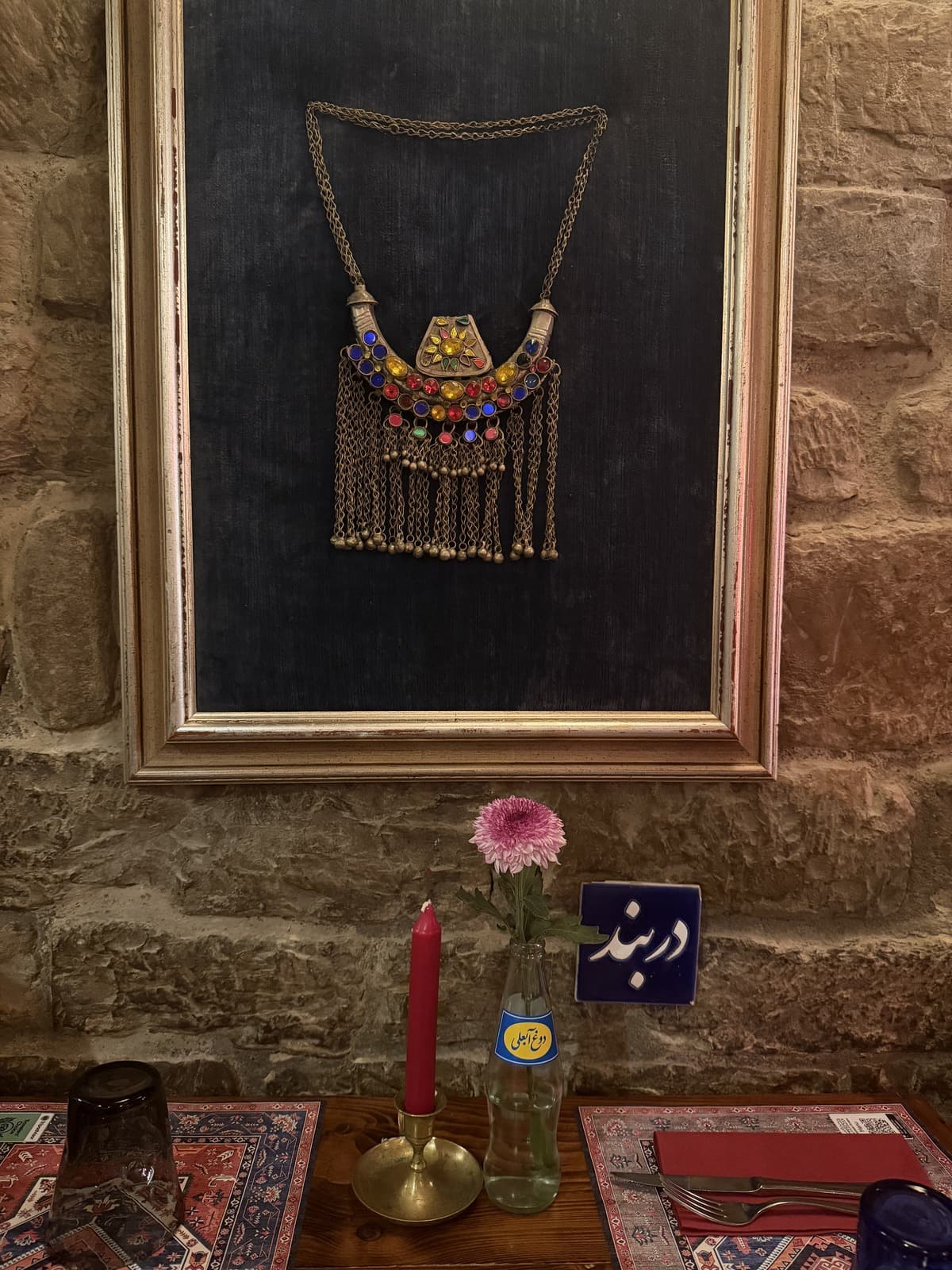 A framed Persian necklace on a stone wall above a candle, a pink flower and a small blue sign